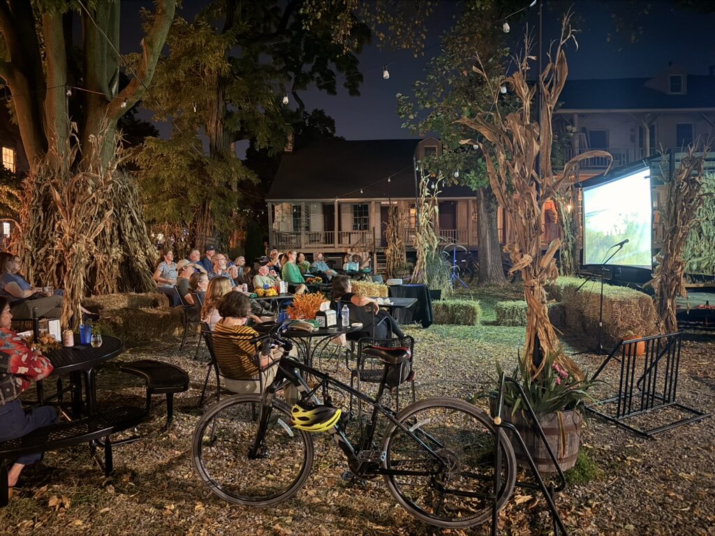 An outdoor screening at night: tables and hay bales among cornstalks, a bicycle in the foreground, the film on a screen under the trees