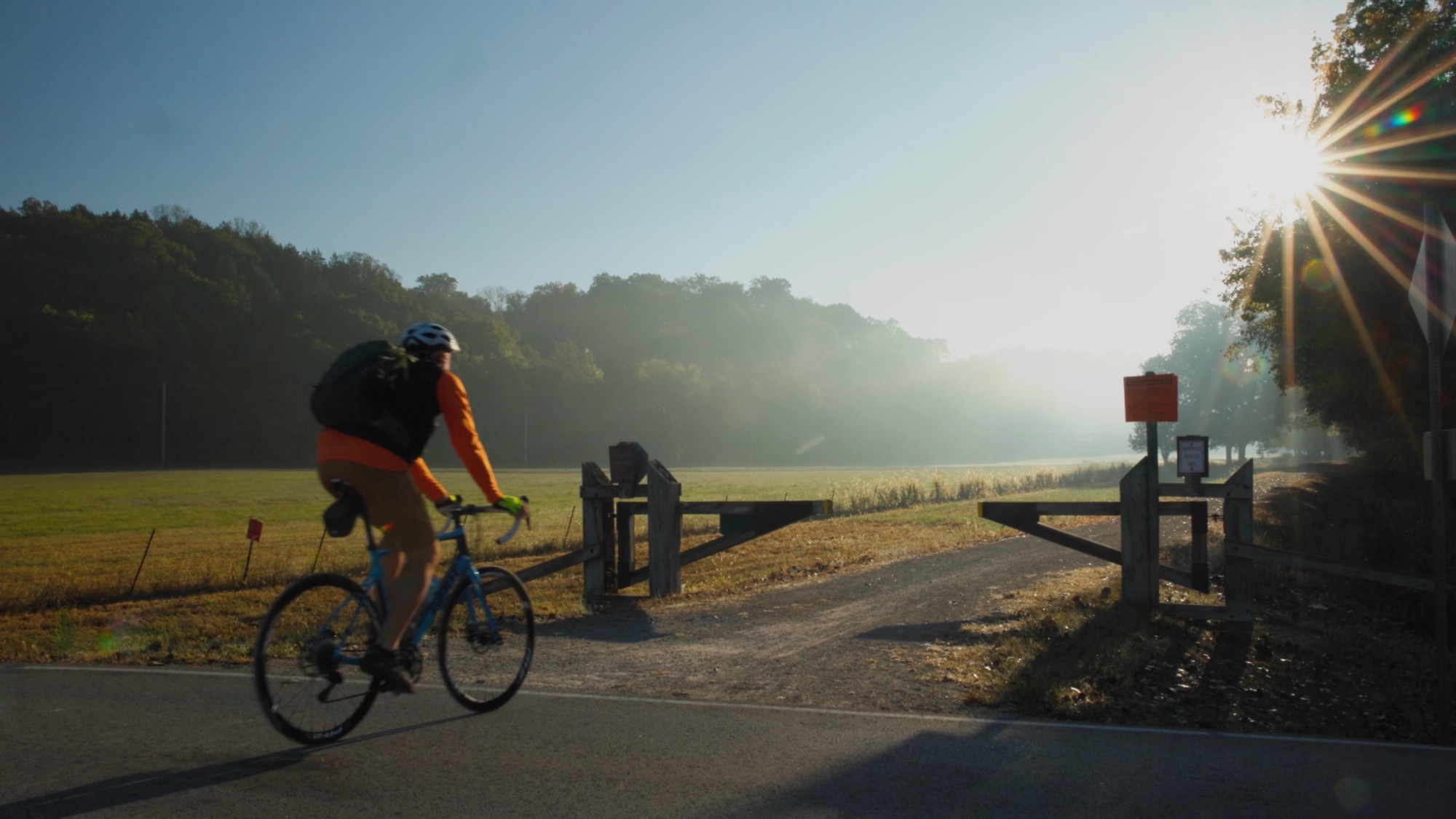A cyclist crossing a road at a Katy Trail trailhead, low sun behind the trees